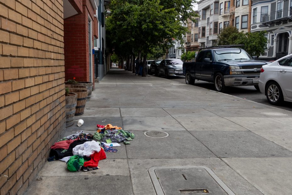 A small pile of assorted clothes and a cup are scattered on the sidewalk next to a brick building on a quiet urban street with parked cars and trees.