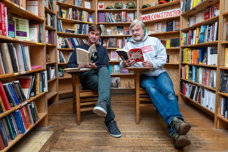 Two men sit on chairs in a bookstore, reading books surrounded by shelves filled with various books; a "Under Construction" sign hangs above.