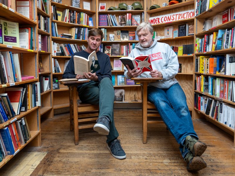 Two men sit on chairs in a bookstore, reading books surrounded by shelves filled with various books; a "Under Construction" sign hangs above.