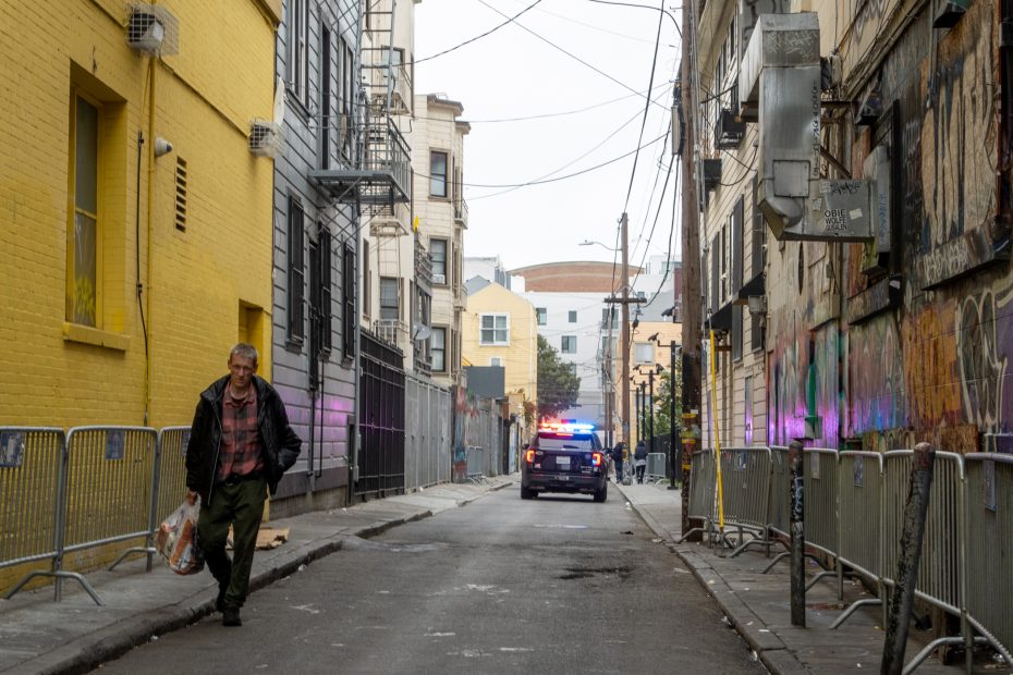 A man carrying a plastic bag walks down an urban alley as a police car with flashing lights is parked ahead. The walls are covered in graffiti, and metal barriers line the street.