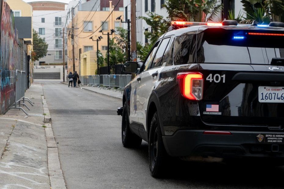 A police SUV with flashing lights is parked on the side of a city alley, with two people visible in the distance near the end of the street.