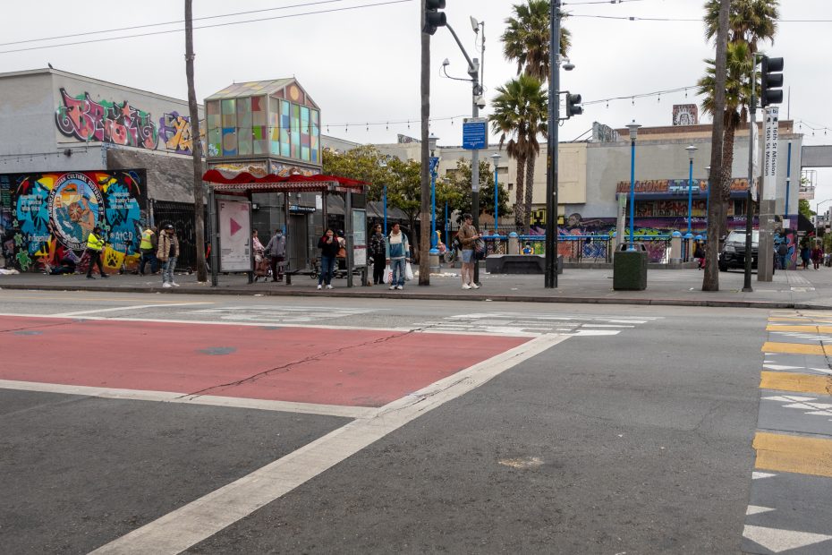 A city street scene shows people waiting at a bus stop with colorful graffiti covering nearby buildings and a crosswalk in the foreground.