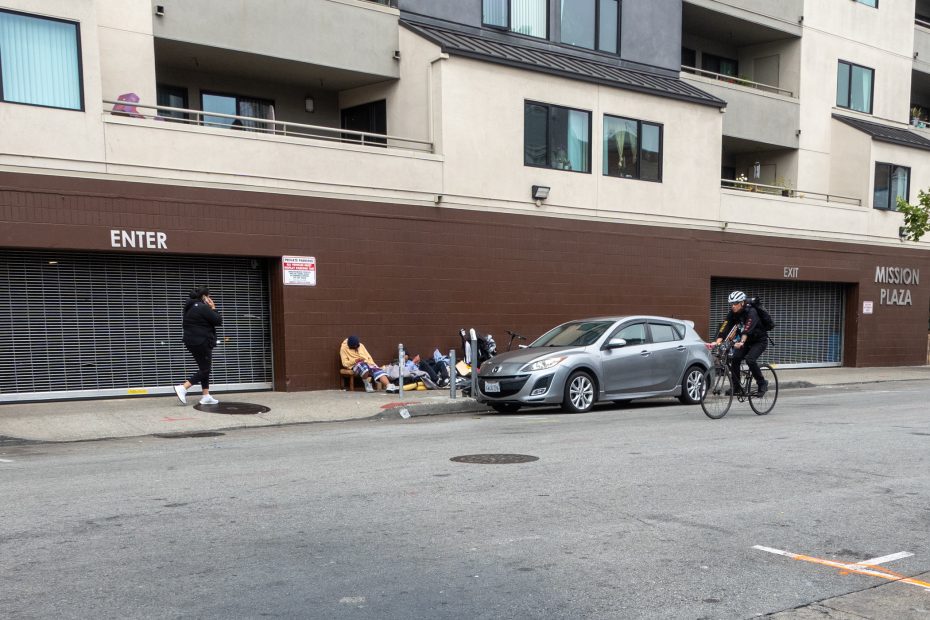 A group of people sit with belongings on a sidewalk outside an apartment building; a cyclist and a pedestrian pass by on the street.