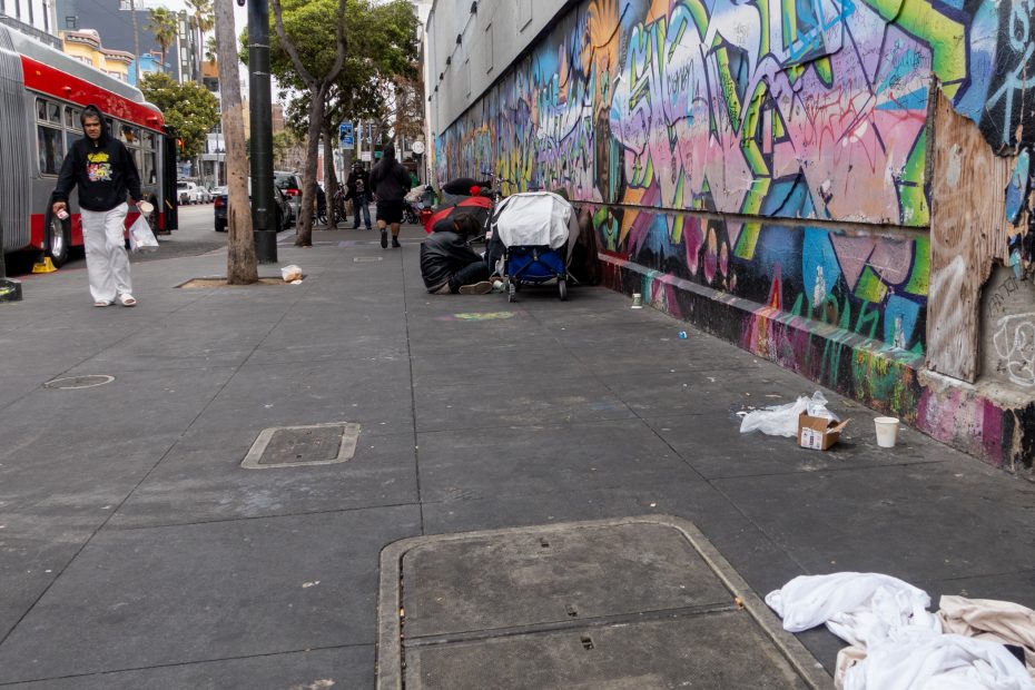 People are gathered with belongings near a colorful graffiti-covered wall on a city sidewalk, with litter and a bus visible in the background.