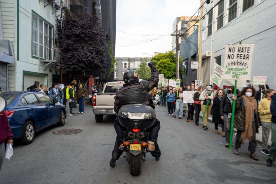 A person on a motorcycle raises a fist while riding through a street crowded with protesters holding signs, some reading “No Hate, No Fear” and “All Are Welcome Here.”.