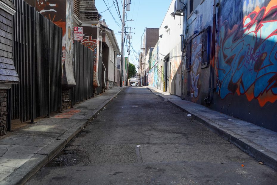 Narrow urban alleyway with graffiti on the right wall, fences and utility poles on the left, and buildings lining both sides under a clear sky.
