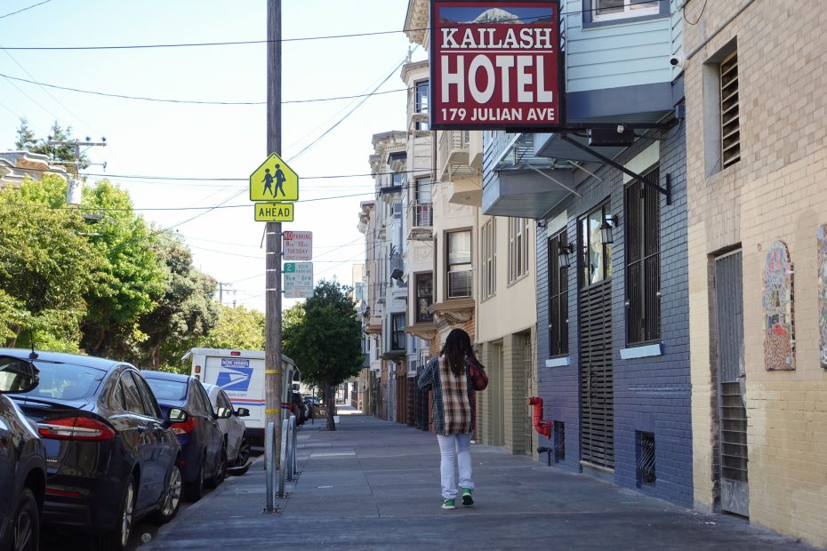A person walks on a sidewalk next to parked cars and buildings, passing a sign for Kailash Hotel at 179 Julian Ave. A pedestrian crossing sign is also visible.