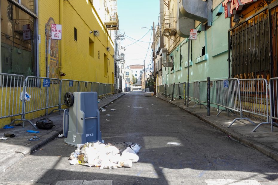 A deserted urban alleyway with metal barricades along both sides, a tipped-over trash can, and scattered litter on the ground.