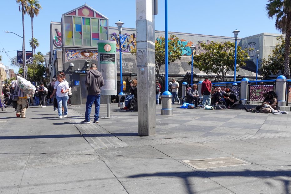 People are gathered on a city sidewalk near a colorful building with murals, some sitting and some standing, on a sunny day.
