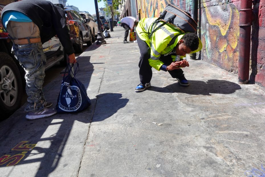 Two men stand on a city sidewalk; one squats down inspecting the ground while the other, holding a large bag, faces away near parked cars and graffiti-covered walls.
