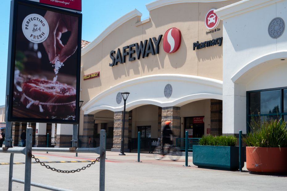 The entrance of a Safeway grocery store with a pharmacy, outdoor signage, and a person riding a bicycle in front.