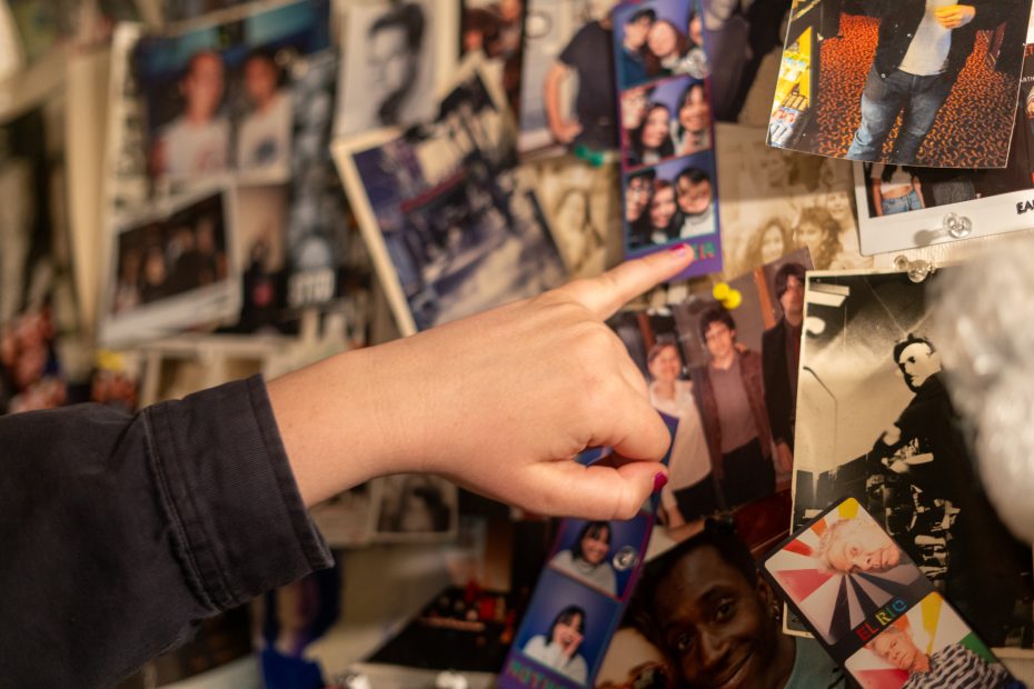 A person's hand points at a collage of various photographs and photo booth strips pinned to a crowded wall.