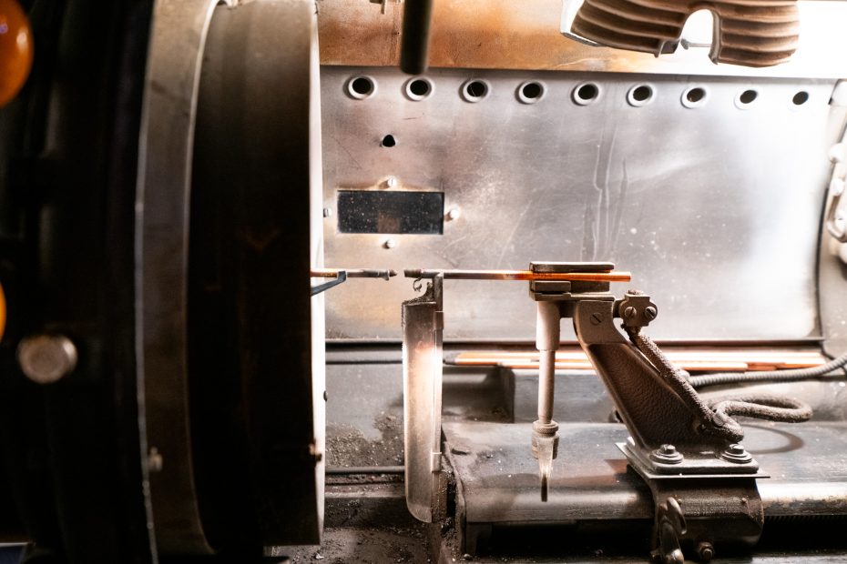 Close-up of a machine tool in a workshop, showing metal rods being processed or measured with various mechanical components and fixtures visible.