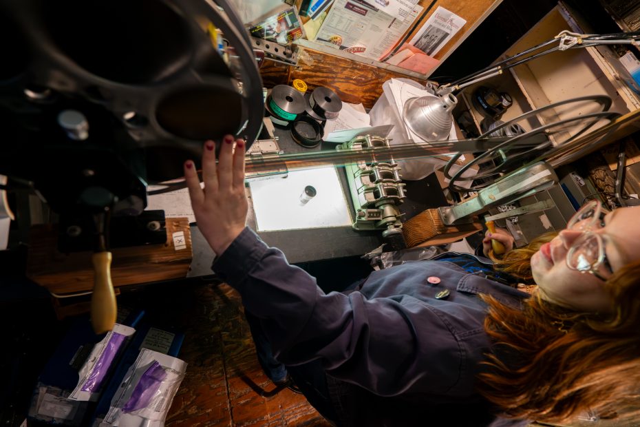 A person examines and handles film reels on a light table in a workspace filled with film equipment and papers.