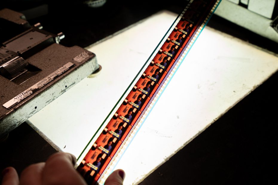 A hand holds a strip of 35mm film featuring multiple frames of an animated orange car, positioned over a lightbox for viewing.