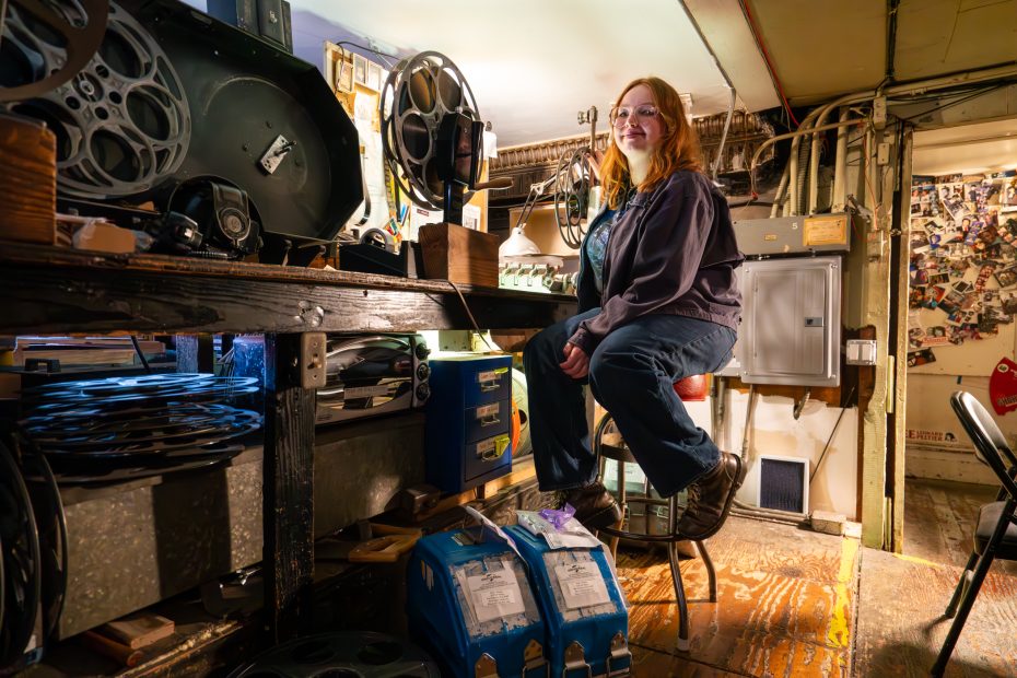 A woman sits on a stool in a film projection room, surrounded by film reels, equipment, and cluttered shelves.