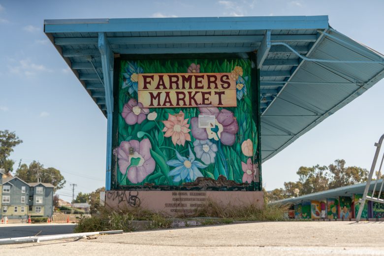 A colorful mural with flowers and a "Farmers Market" sign on a concrete structure under a blue metal roof, with trees and buildings in the background.