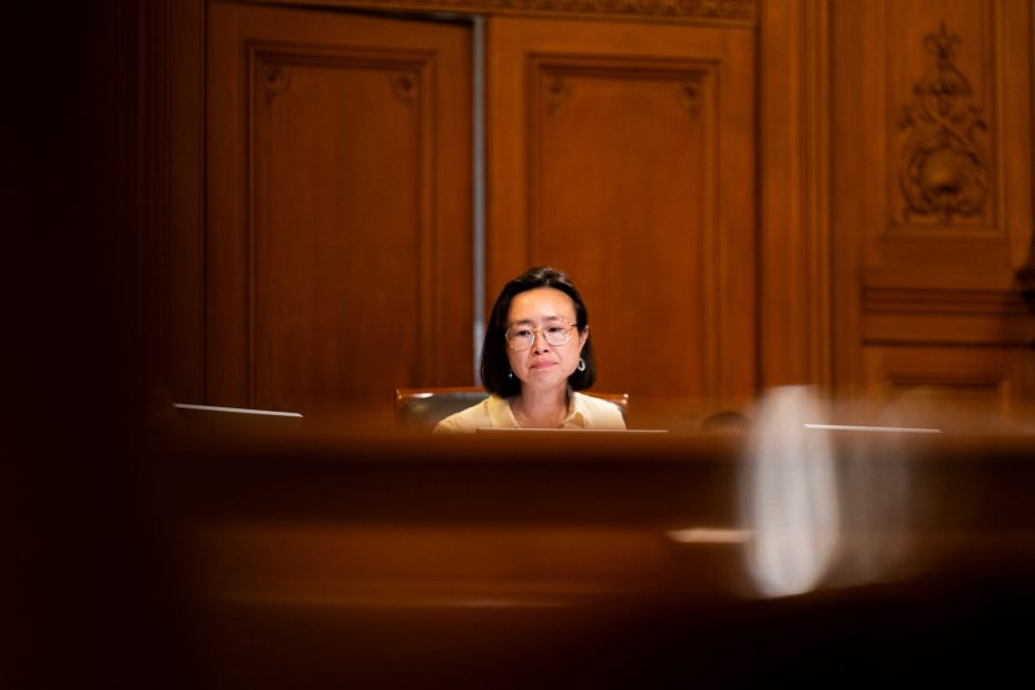 A woman wearing glasses sits at a desk in a wood-paneled room, facing forward with a neutral expression.