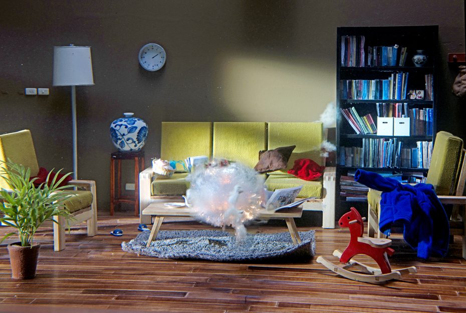 A modern living room with chairs, a sofa, a bookshelf, and a coffee table. Smoke and sparks are emerging from a laptop on the table, suggesting it has exploded or caught fire.