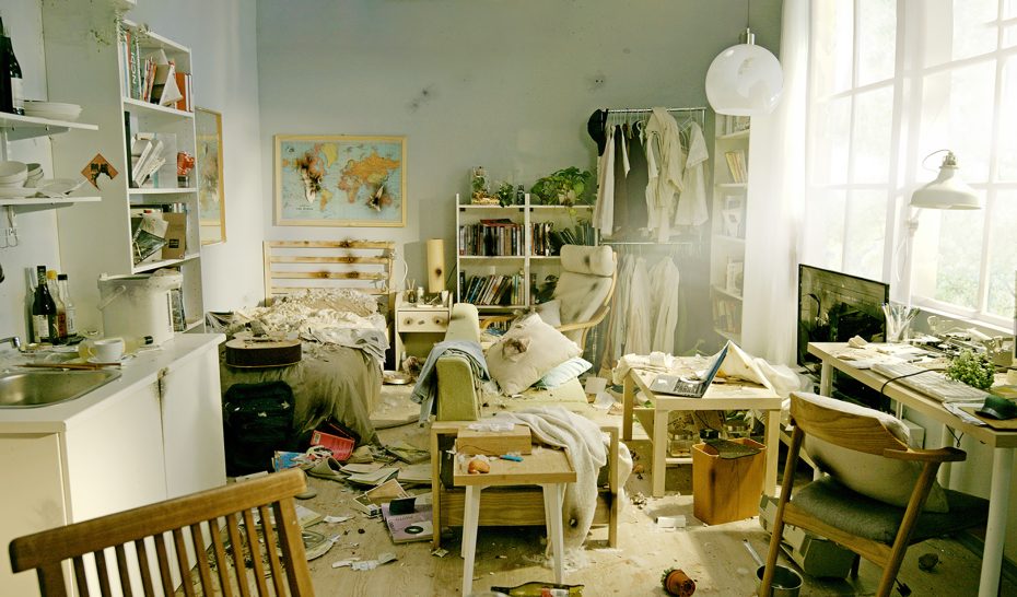 Messy bedroom and living area with scattered clothes, books, and household items, unmade bed, dirty dishes, and sunlight streaming through large windows.