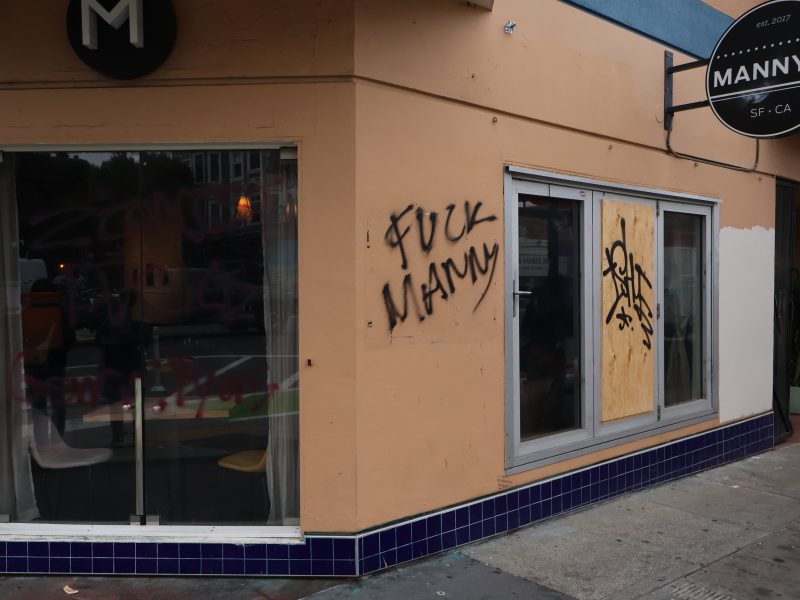 Graffiti and vandalism on the exterior of Manny's storefront, including spray-painted messages and boarded-up windows.