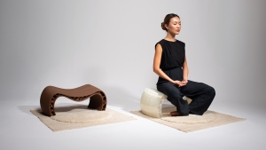 A woman sits cross-legged on a translucent, curved seat, with a brown, similarly shaped seat placed nearby on a separate rug.