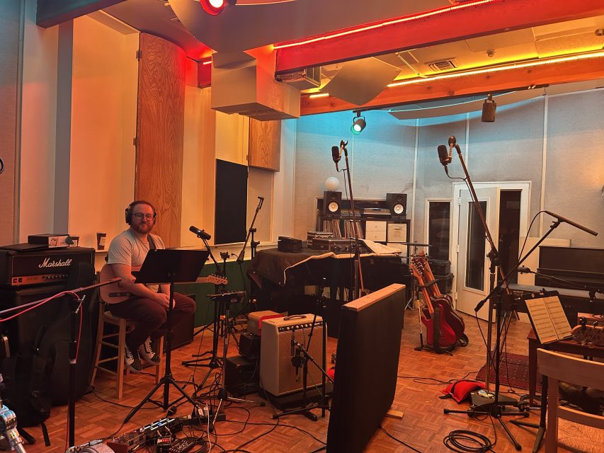 A man sits on a stool in a recording studio surrounded by musical equipment, microphones, amplifiers, and guitars.