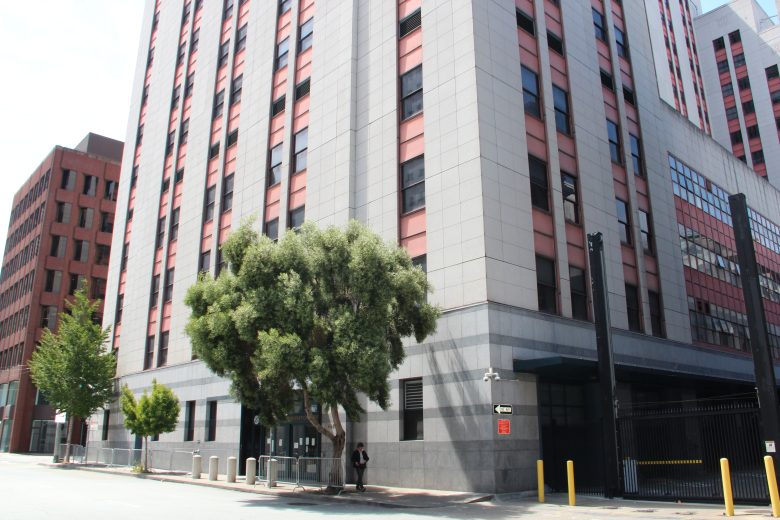 Tall modern office building with gray stone facade and red vertical window panels, a large tree in front, and a gated entrance on the right.