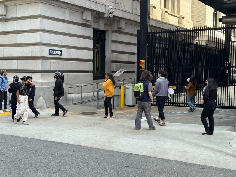 A group of people walks and stands near a gated entrance of a large building on a city street corner; some hold papers or signs.