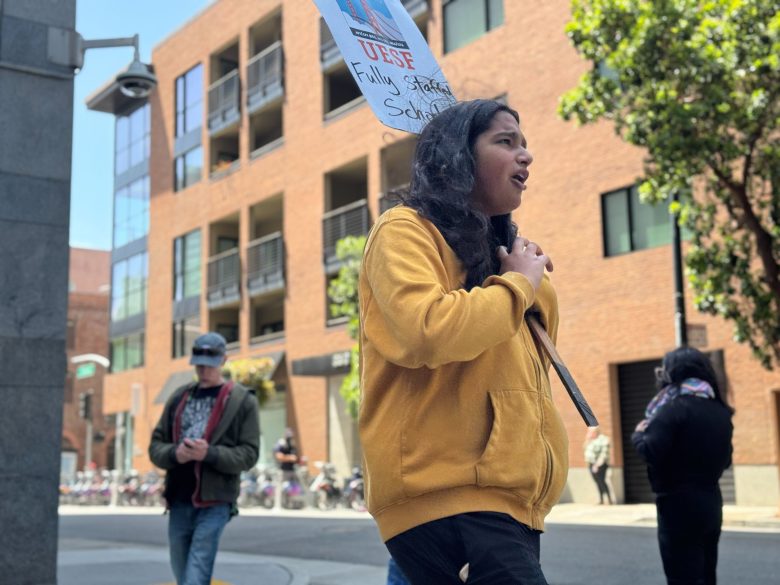 A person wearing a yellow hoodie holds a protest sign while walking on a city street; other people and brick buildings are visible in the background.