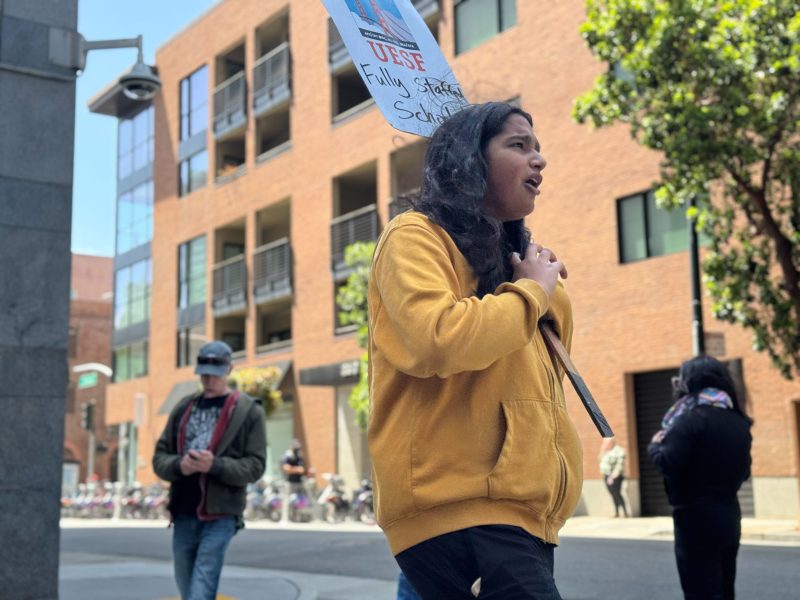 A person wearing a yellow hoodie holds a protest sign while walking on a city street; other people and brick buildings are visible in the background.