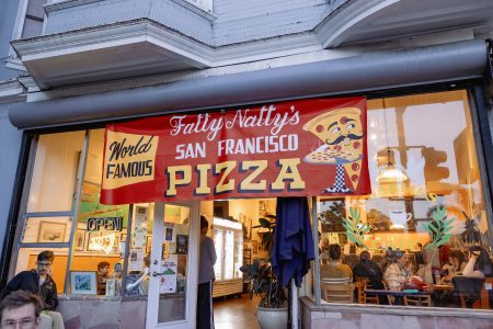 A banner above a pizzeria reads "Fatty Natty's World Famous San Francisco Pizza"; people are seated inside and outside the restaurant.