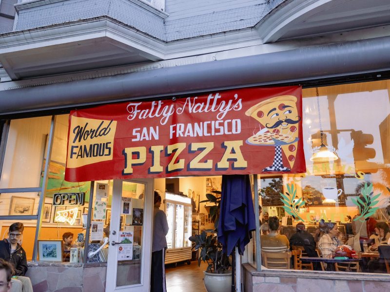 A banner above a pizzeria reads "Fatty Natty's World Famous San Francisco Pizza"; people are seated inside and outside the restaurant.