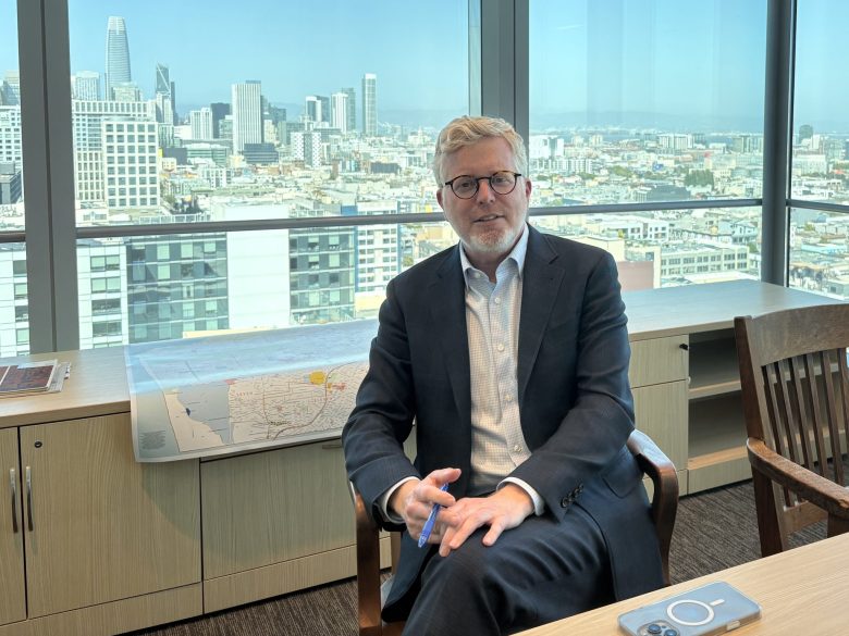 A man in a suit sits in a modern office with large windows overlooking a city skyline. There are papers and a laptop on the desk nearby.