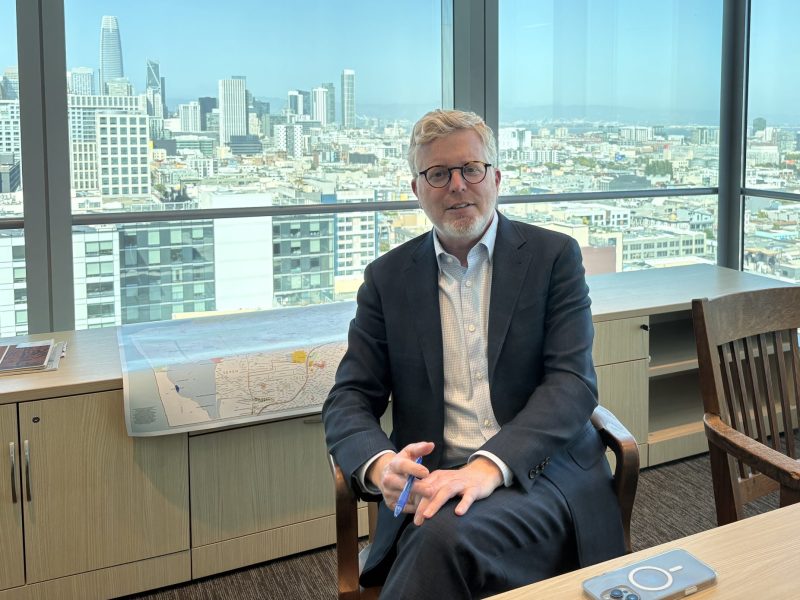 A man in a suit sits in a modern office with large windows overlooking a city skyline. There are papers and a laptop on the desk nearby.