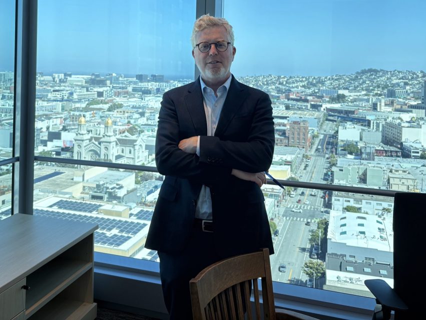 A man in a suit stands with arms crossed in a modern office with large windows overlooking a cityscape on a clear day.