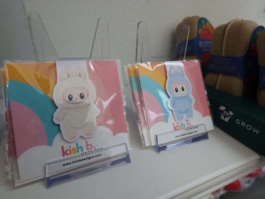 Two clear display stands hold cards with cartoon characters in animal onesies, one in white and one in blue, labeled "kish b." Next to them are seed starter packs.
