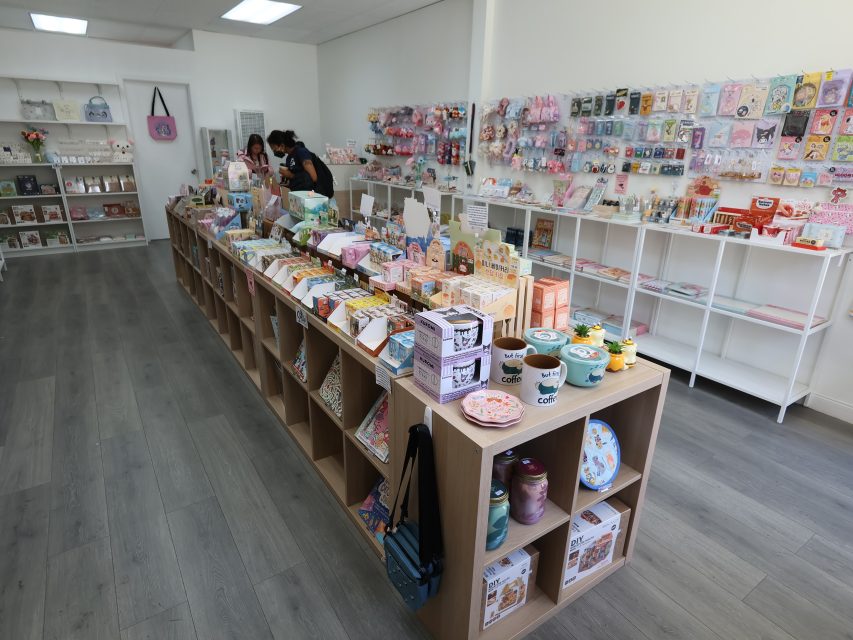 A store interior with shelves and tables displaying various colorful stationery, toys, and gift items; two people stand near the back counter.