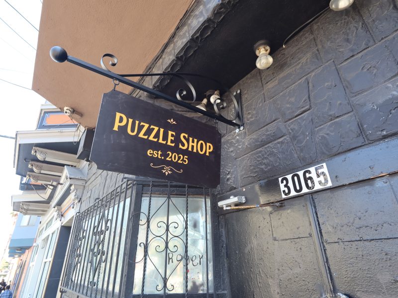A black and gold sign reading "Puzzle Shop est. 2025" hangs above a doorway next to an address labeled 3065 on a dark brick building.