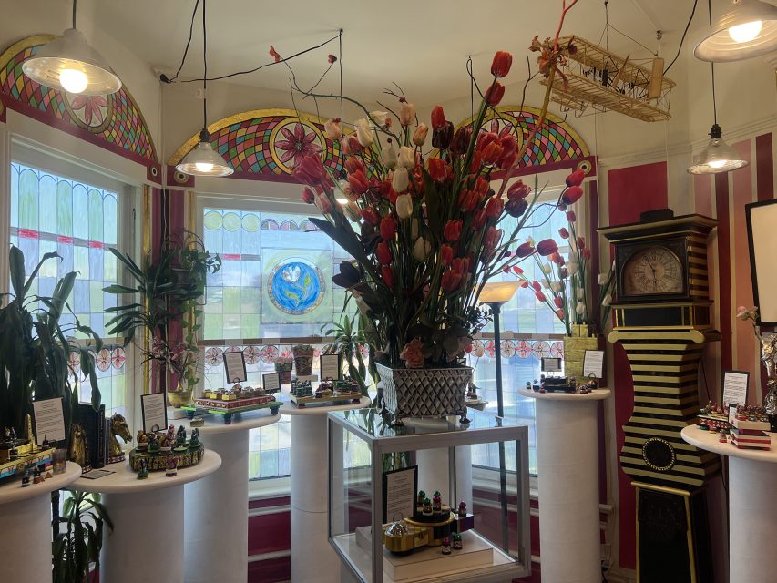 A room with colorful stained-glass windows, a large floral arrangement on a pedestal table, plants, an ornate clock, and small decorated items on display.