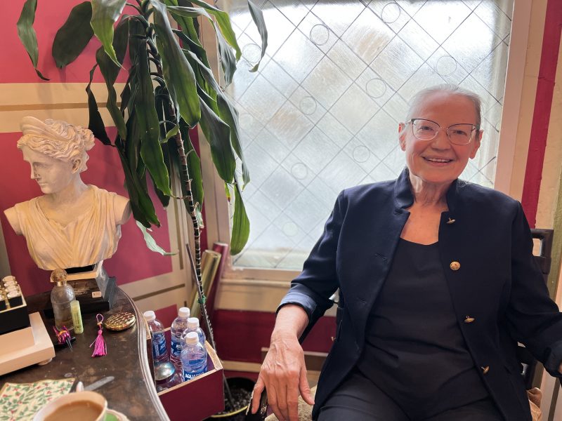 An older woman with short gray hair and glasses sits smiling beside a desk with a bust, plants, bottled water, and a coffee cup, in a brightly lit room with a window.