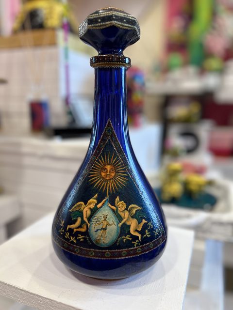 A blue glass bottle with gold accents features painted cherubs, a sun with a face, and a small human figure on its front; displayed on a white pedestal indoors.