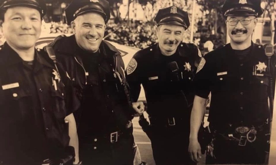 Four uniformed police officers stand together outdoors, smiling for the camera in a black-and-white photograph.
