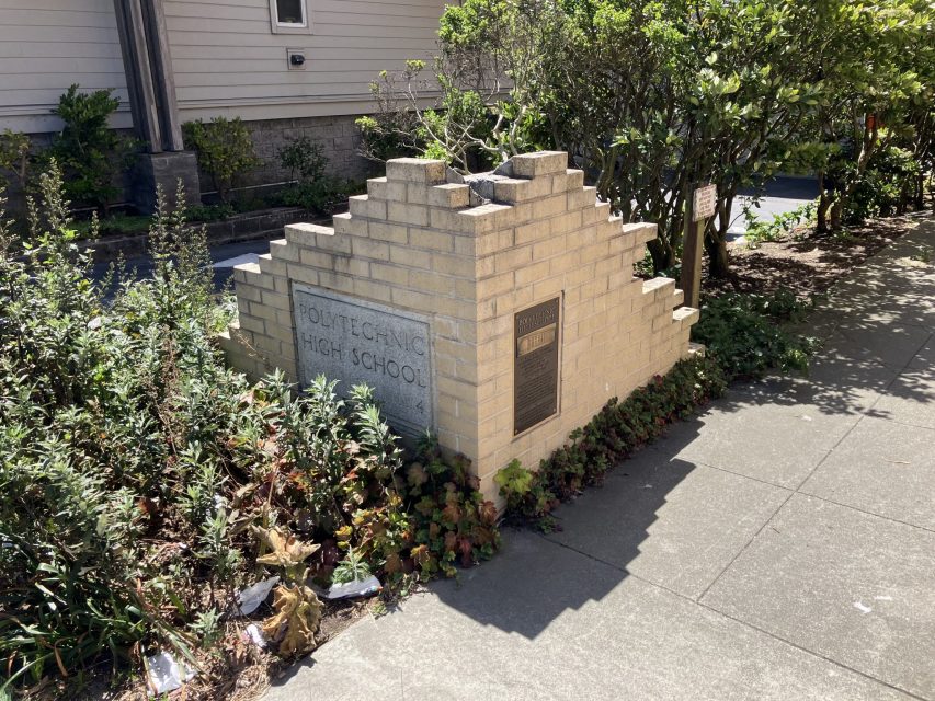 A brick monument with a plaque and a stone inscribed "Polytechnic High School" stands beside a sidewalk, surrounded by plants and bushes.