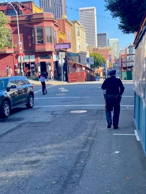 A person walks on a city sidewalk toward an intersection with cars parked and buildings in the background on a sunny day.