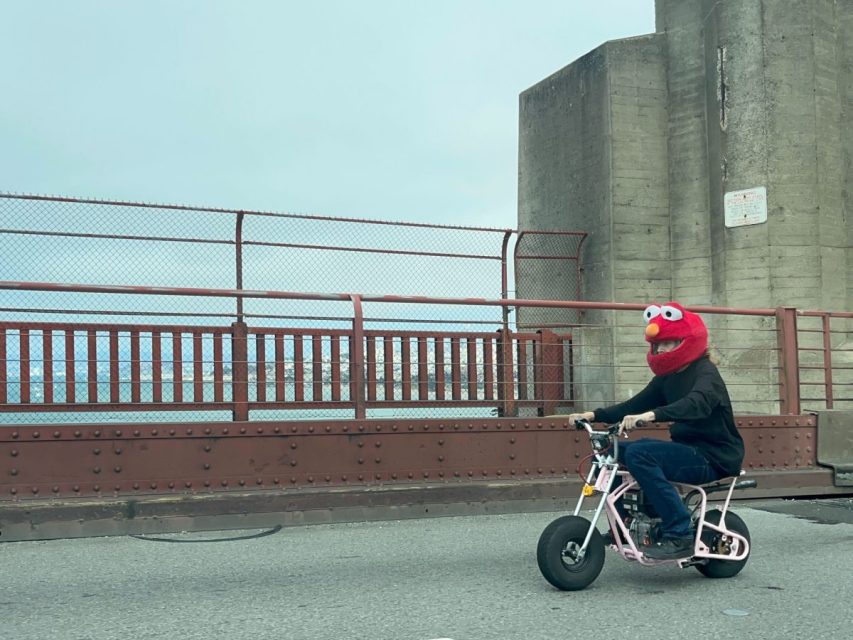 A person wearing an Elmo head costume rides a small motorbike on a bridge with a fence and concrete pillars in the background.
