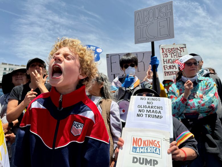 A crowd of protesters holds signs with messages opposing political figures and policies; a child in a USA jacket shouts in the foreground.