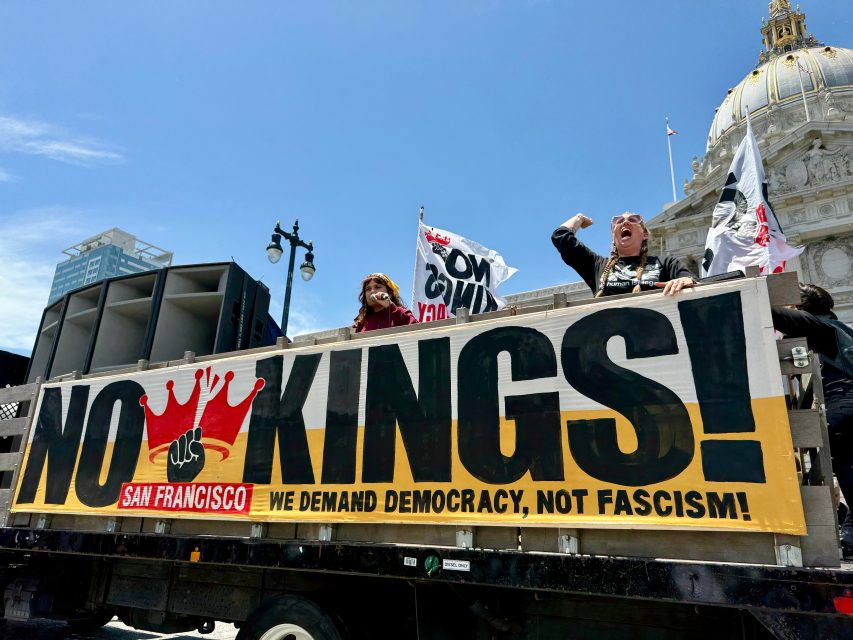 Protesters stand on a truck with a large banner reading "NO KINGS! We demand democracy, not fascism!" in front of a government building in San Francisco.