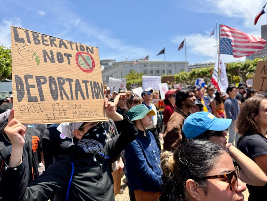A crowd at a protest holds signs, including one reading "Liberation Not Deportation" with smaller text "Free Kilmar, Free Mahoud"; American flags are visible in the background.