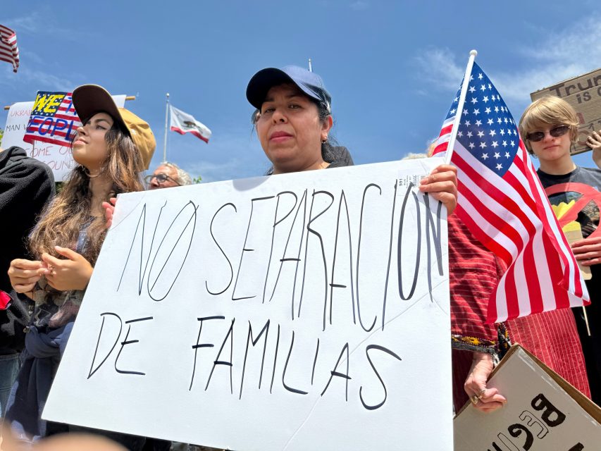 A woman holds a sign reading "No Separacion de Familias" at a protest, surrounded by other people, some holding American flags.